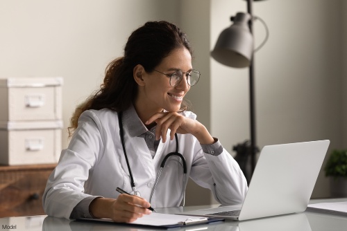 Physician smiling at a laptop and taking notes on a clipboard